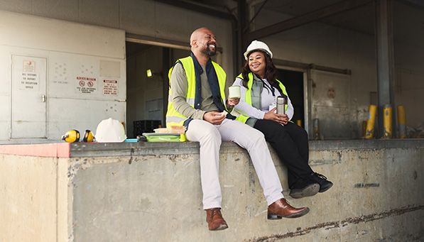 Two workers in reflective vests sit on a loading dock, holding drinks during a break.