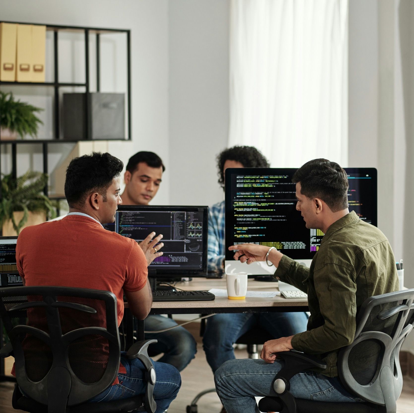 Four colleagues work together at computer monitors displaying code in a sunlit office.
