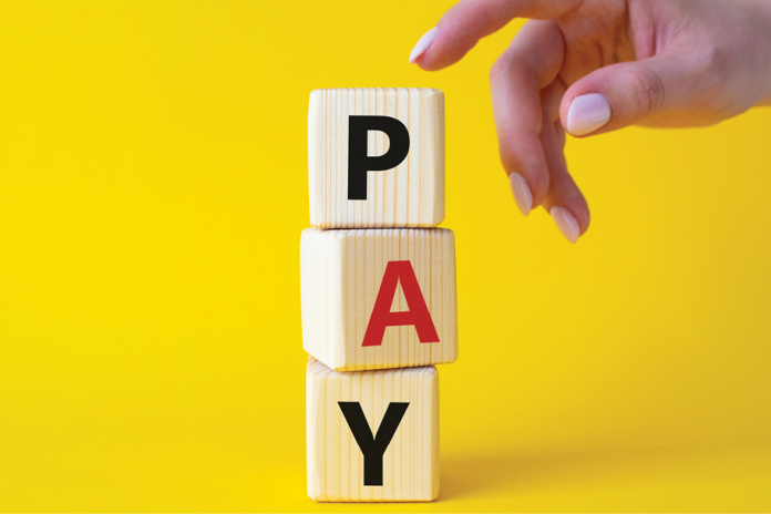 A hand reaches toward stacked wooden blocks spelling Pay on a yellow background.