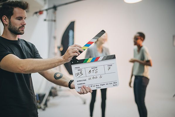 A crew member holds an open clapperboard on a film set with people behind him.