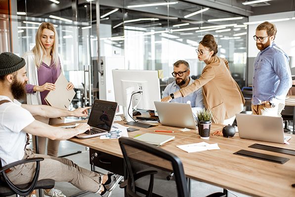 Five coworkers collaborate around computers at a shared office desk, including a wheelchair user.