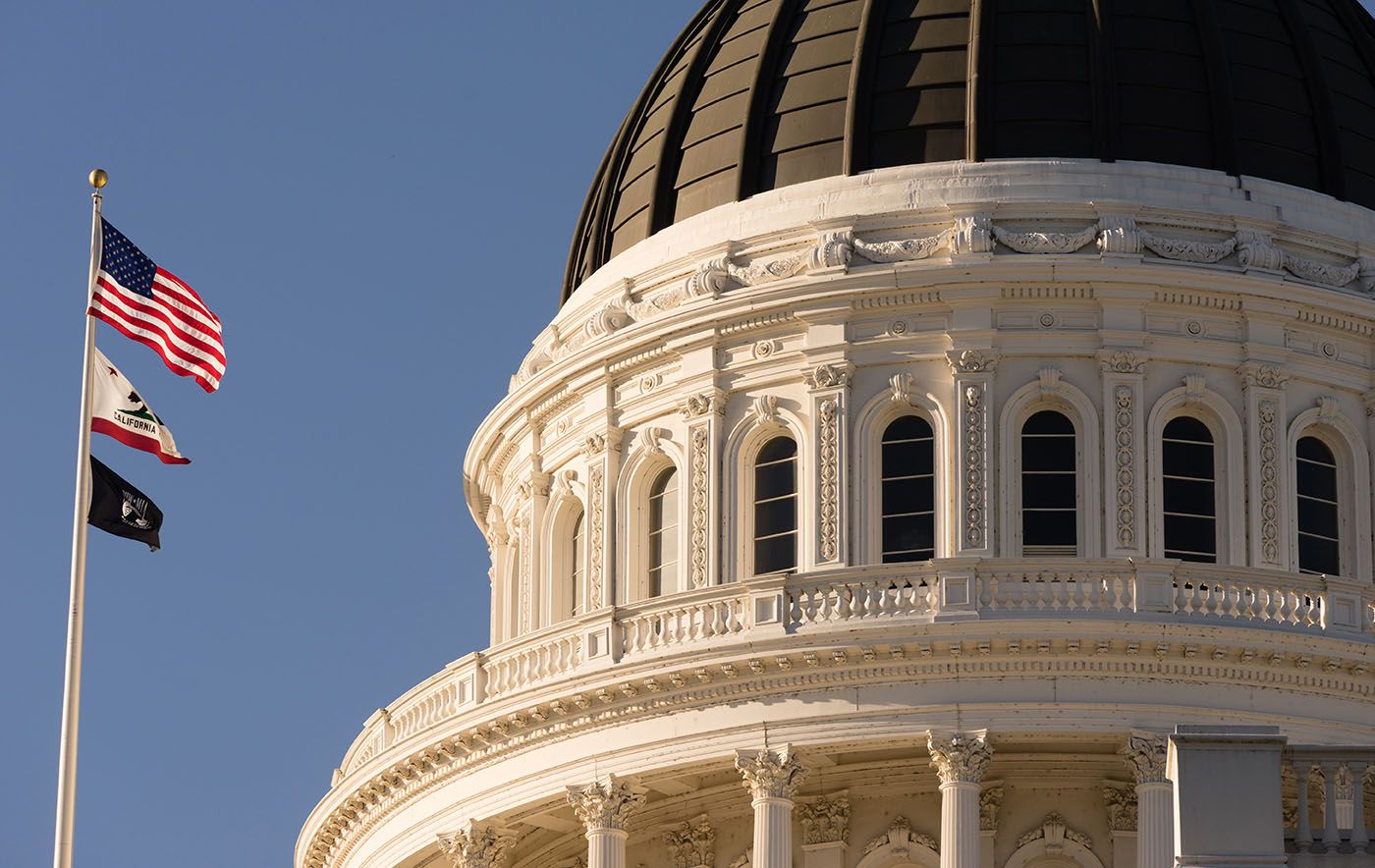 California State Capitol dome beneath a clear blue sky with United States and California flags.