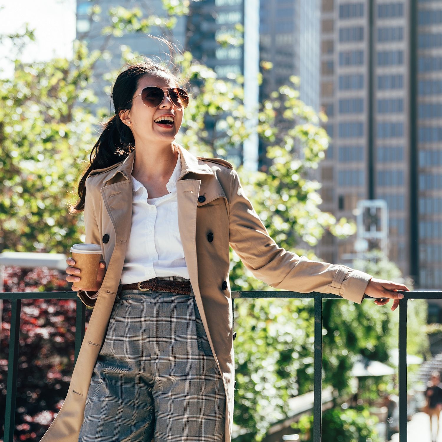 A woman wearing sunglasses stands by a railing in a city plaza holding a takeaway cup.