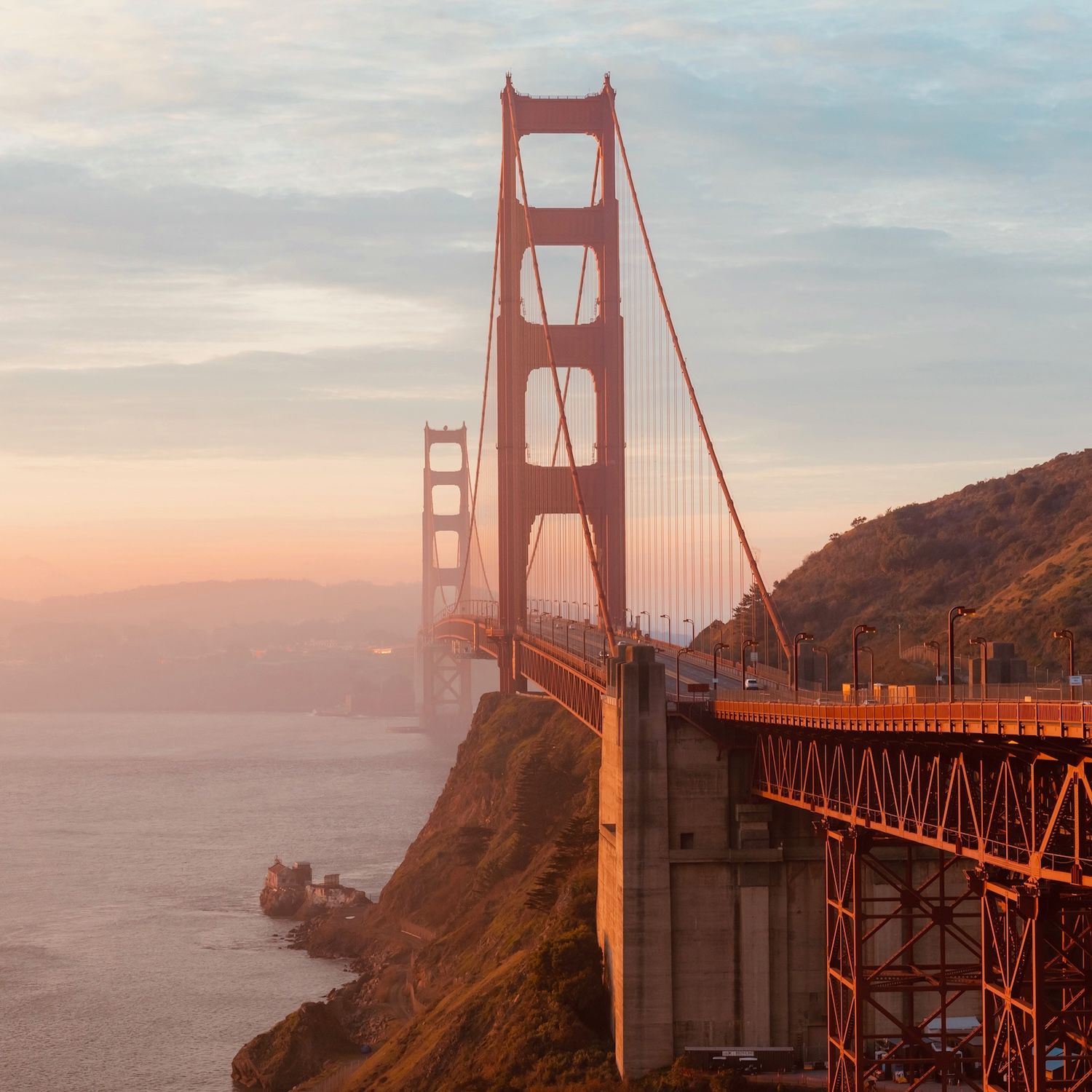 The Golden Gate Bridge stretches over foggy water in warm evening light.
