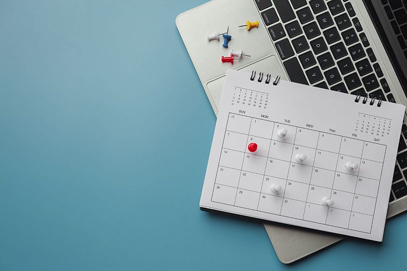 A paper calendar and colorful pushpins lie on a laptop on a blue surface.