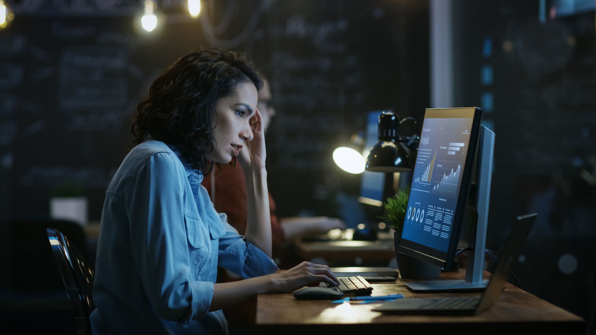 A woman works at a desktop computer in a dimly lit office at night.