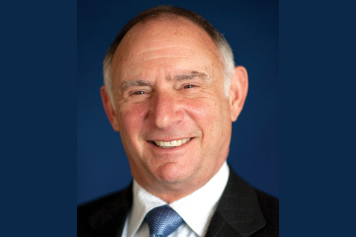 Portrait of a smiling man wearing a dark suit and blue tie against a blue background.