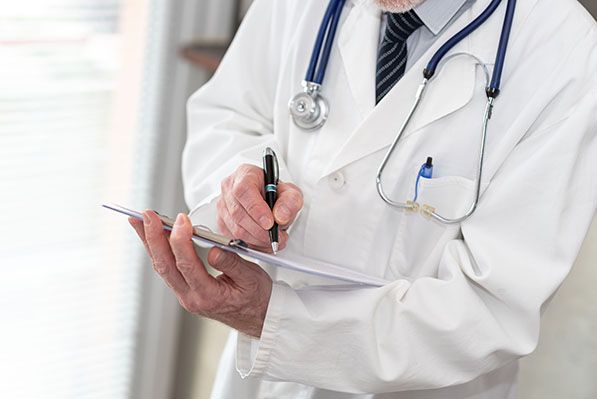 A clinician wearing a white coat and stethoscope writes on a clipboard.