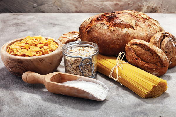 Bread loaves, pasta, oats, cornflakes and flour arranged on a stone surface.