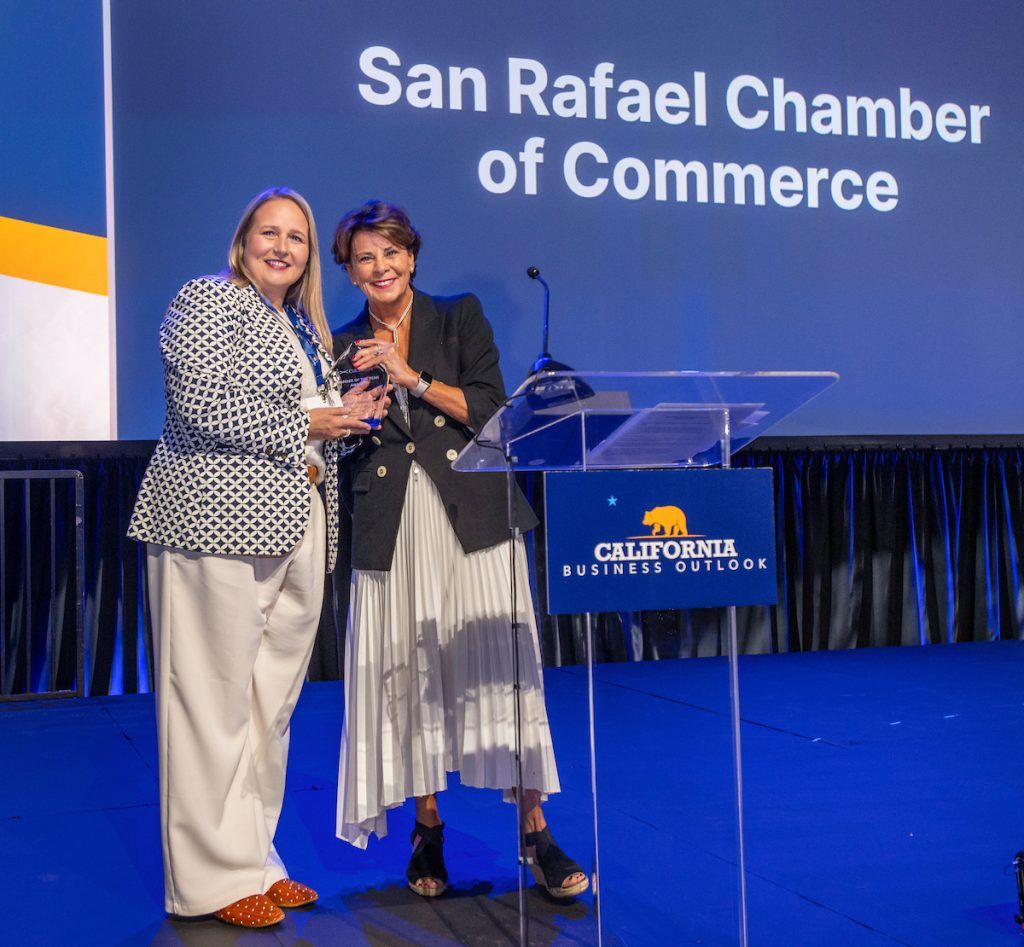 Two women hold an award beside a lectern and a screen reading San Rafael Chamber of Commerce.