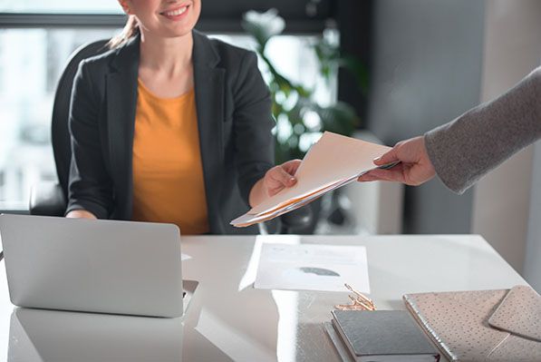 A person hands a folder to an employee seated at a desk with a laptop.