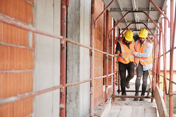 Two construction workers in hard hats and safety vests walk together through scaffolding.