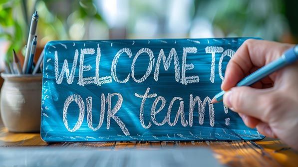 A hand writes WELCOME TO OUR team on a small blue chalkboard.
