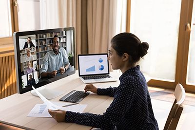 A woman joins a video call at a desk with a desktop monitor and a laptop showing charts.