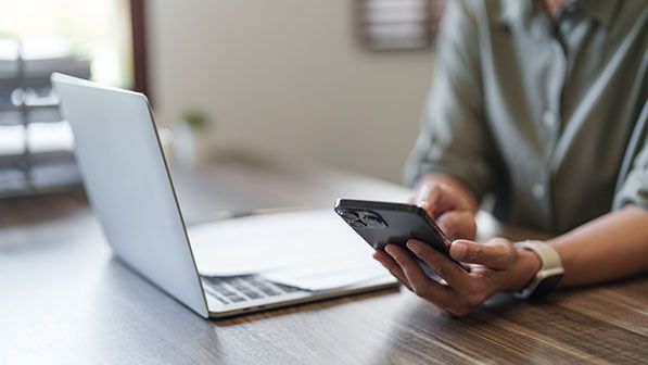 A person uses a smartphone while a laptop sits open on a desk.
