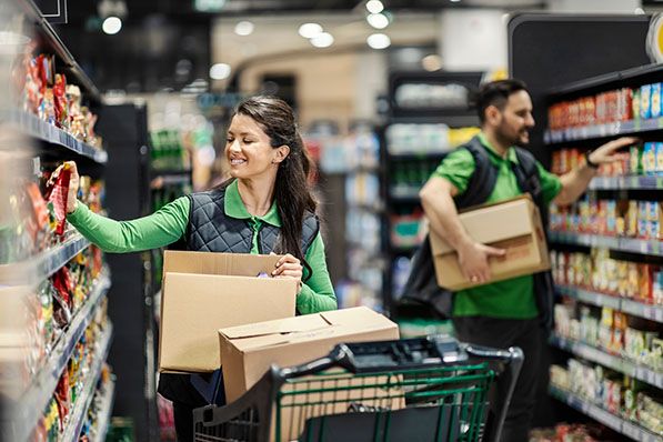 Two grocery workers restock shelves while carrying cardboard boxes.