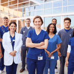 A group of healthcare workers in scrubs and lab coats poses in a bright hospital hallway.