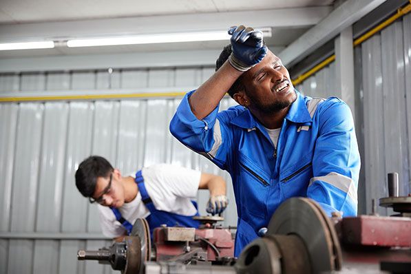 A worker in blue coveralls wipes his forehead in an industrial workshop.