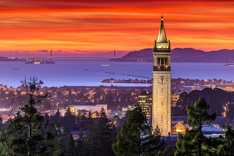 Berkeley’s Campanile tower rises above trees with San Francisco Bay beneath an orange sunset.