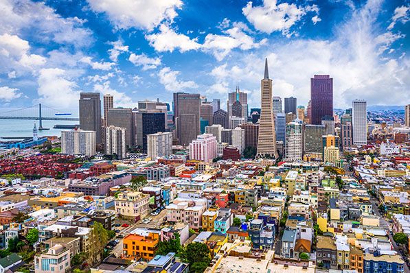 Aerial view of San Francisco’s dense neighborhoods, downtown towers and bay beneath scattered clouds.