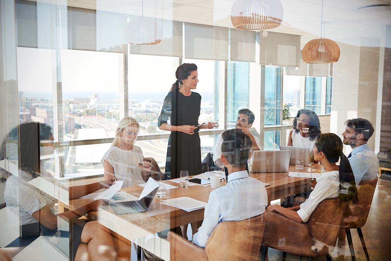 A diverse group of professionals in a modern glass conference room, with a woman presenting to colleagues seated around a table overlooking a city.