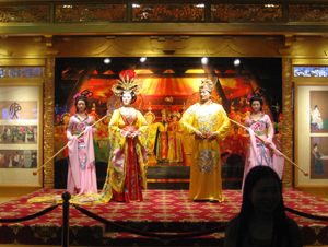 Performers in elaborate colorful robes and headdresses stand on an ornate indoor stage.