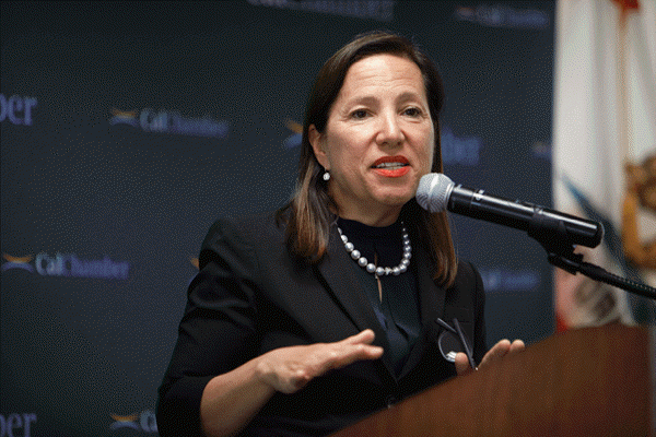 Woman in a dark suit and pearl necklace speaking into a microphone at a podium, with "CalChamber" visible in the background.