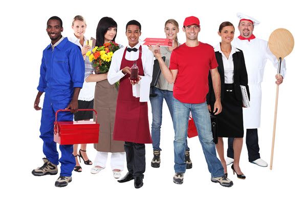 A group of workers in varied uniforms stand together holding tools, flowers, a food tray and a pizza paddle.