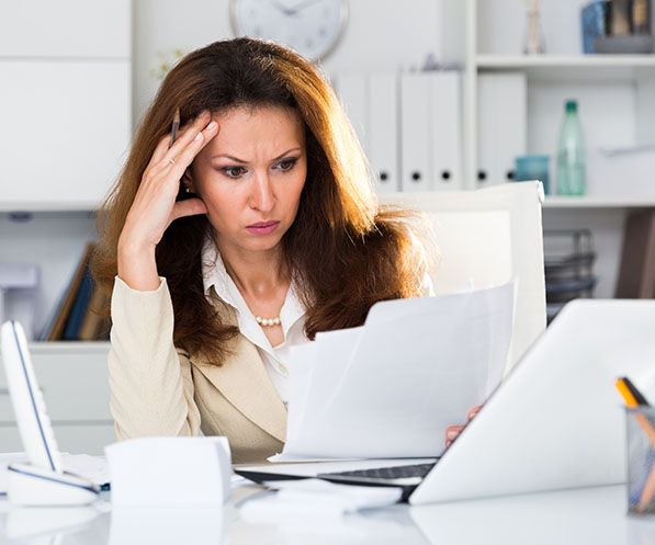 A woman reviews paperwork at a desk while resting a hand against her forehead.