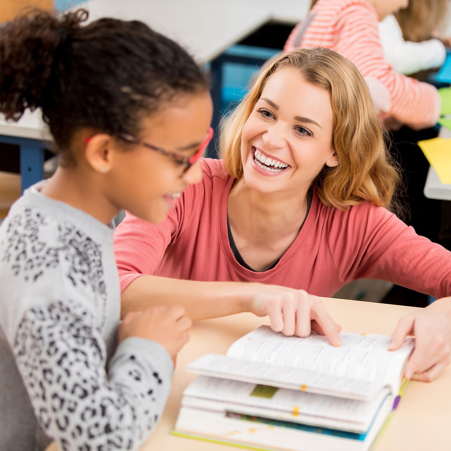A teacher smiles while helping a child with an open workbook.
