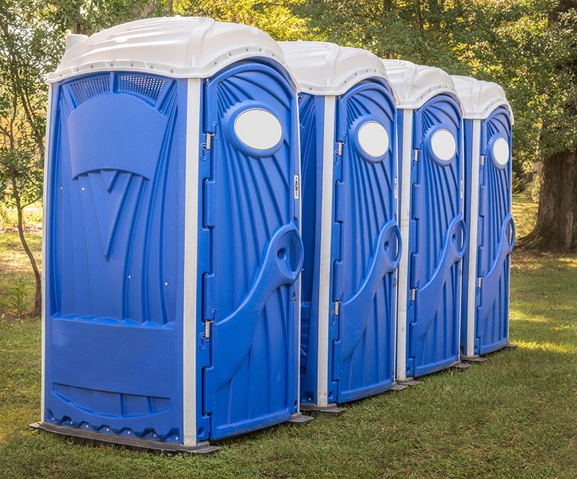 Four blue portable toilets stand in a row on grass beside trees.