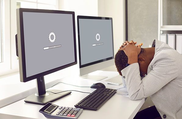 A person leans over a desk holding their head while two monitors display loading indicators.