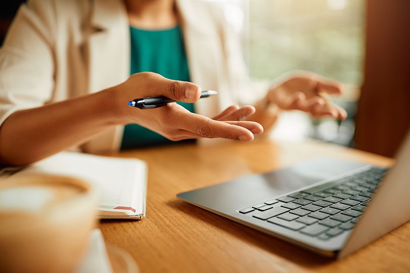 A person gestures with a pen beside an open laptop on a wooden desk.