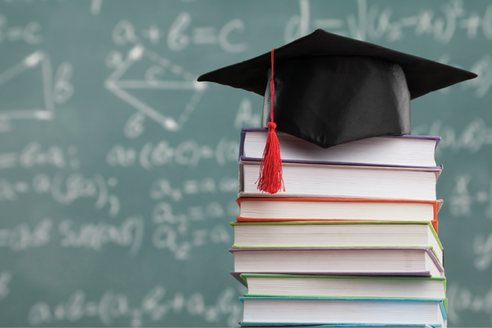A black graduation cap with a red tassel rests on a stack of books before a chalkboard.