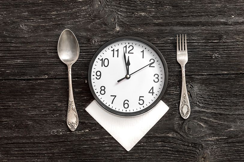 An analog clock on a folded napkin between a spoon and fork on dark wooden boards.