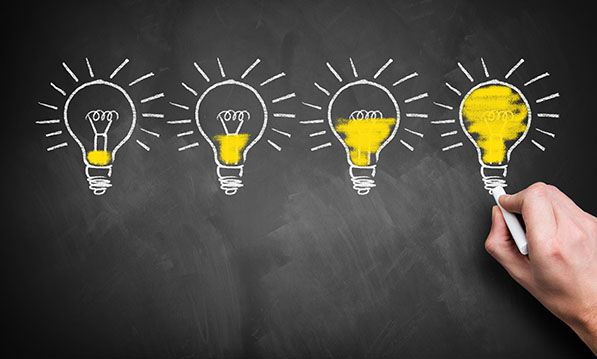 A hand colors the last of four chalk-drawn lightbulbs, each progressively more yellow.