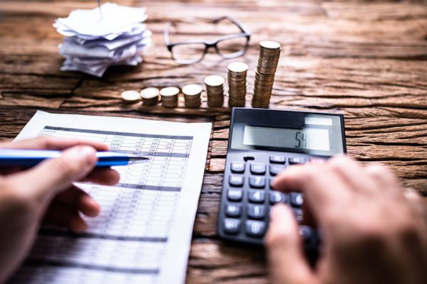 Hands work with a calculator and financial paperwork beside ascending stacks of coins.
