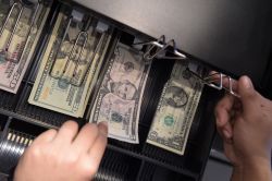Hands sort dollar bills in an open cash register drawer.