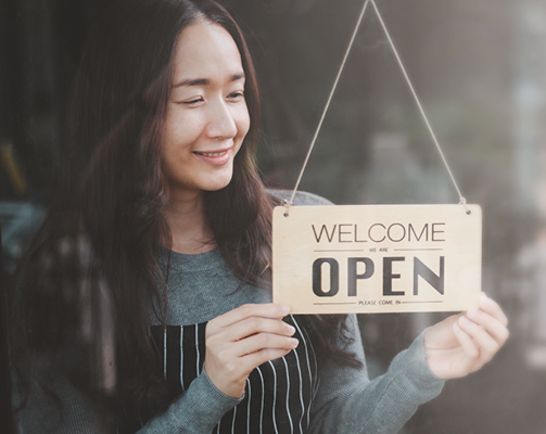 A smiling shop worker holds a Welcome Open sign against a glass door.