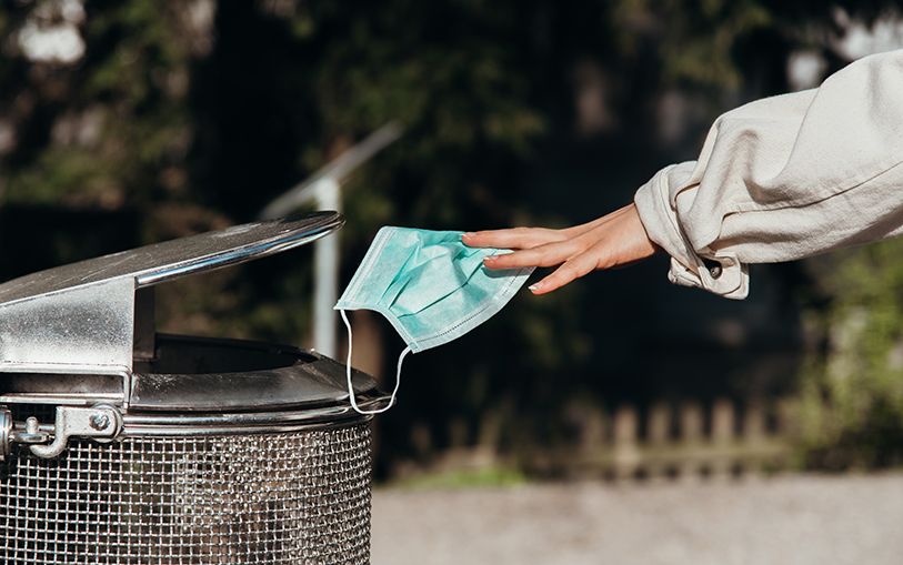 A hand drops a blue disposable face mask into an outdoor trash bin.