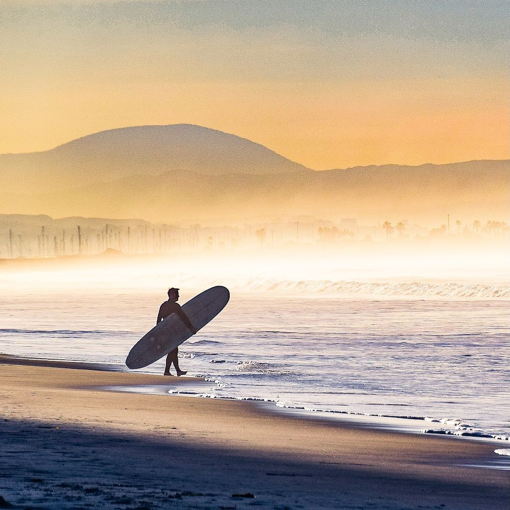 A person carries a surfboard toward the waves on a misty beach at sunrise.
