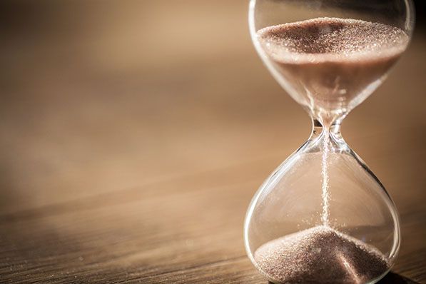 A glass hourglass with sand collecting in its lower bulb stands on a wooden surface.
