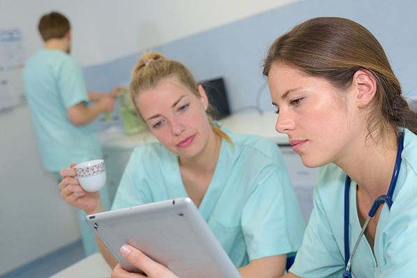 Two healthcare workers in scrubs review a tablet, one holding a small cup.