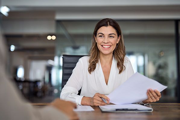Smiling woman in a white shirt holding documents at a desk.