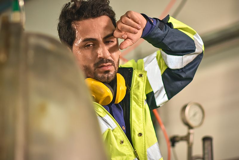 A worker in a reflective vest wipes their forehead with an arm, with ear protectors around their neck.