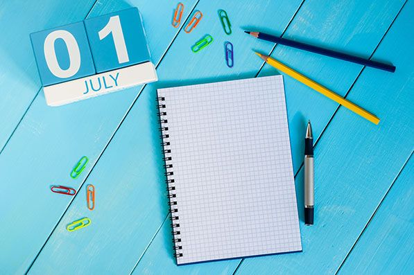 July 1 calendar blocks beside an open notebook, pencils and paper clips on a blue desk.