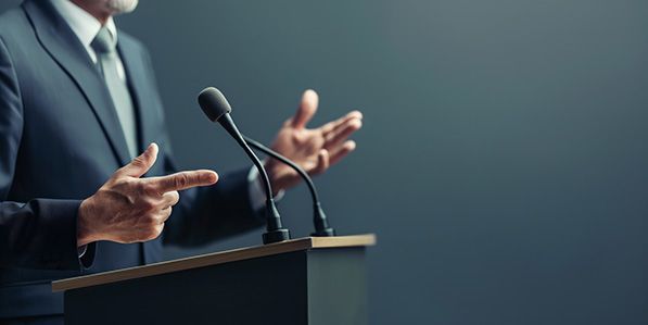 A person in a dark suit gestures with both hands while speaking at a lectern microphone.