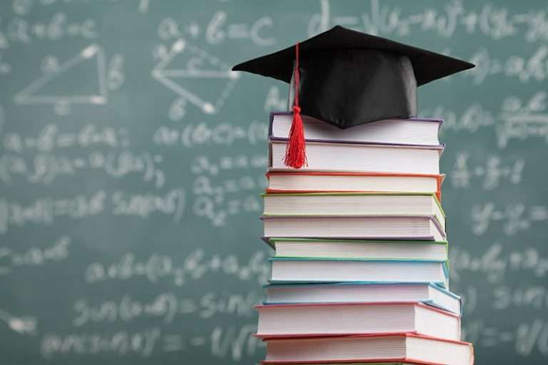 A graduation cap sits atop a stack of books in front of a chalkboard covered in equations.