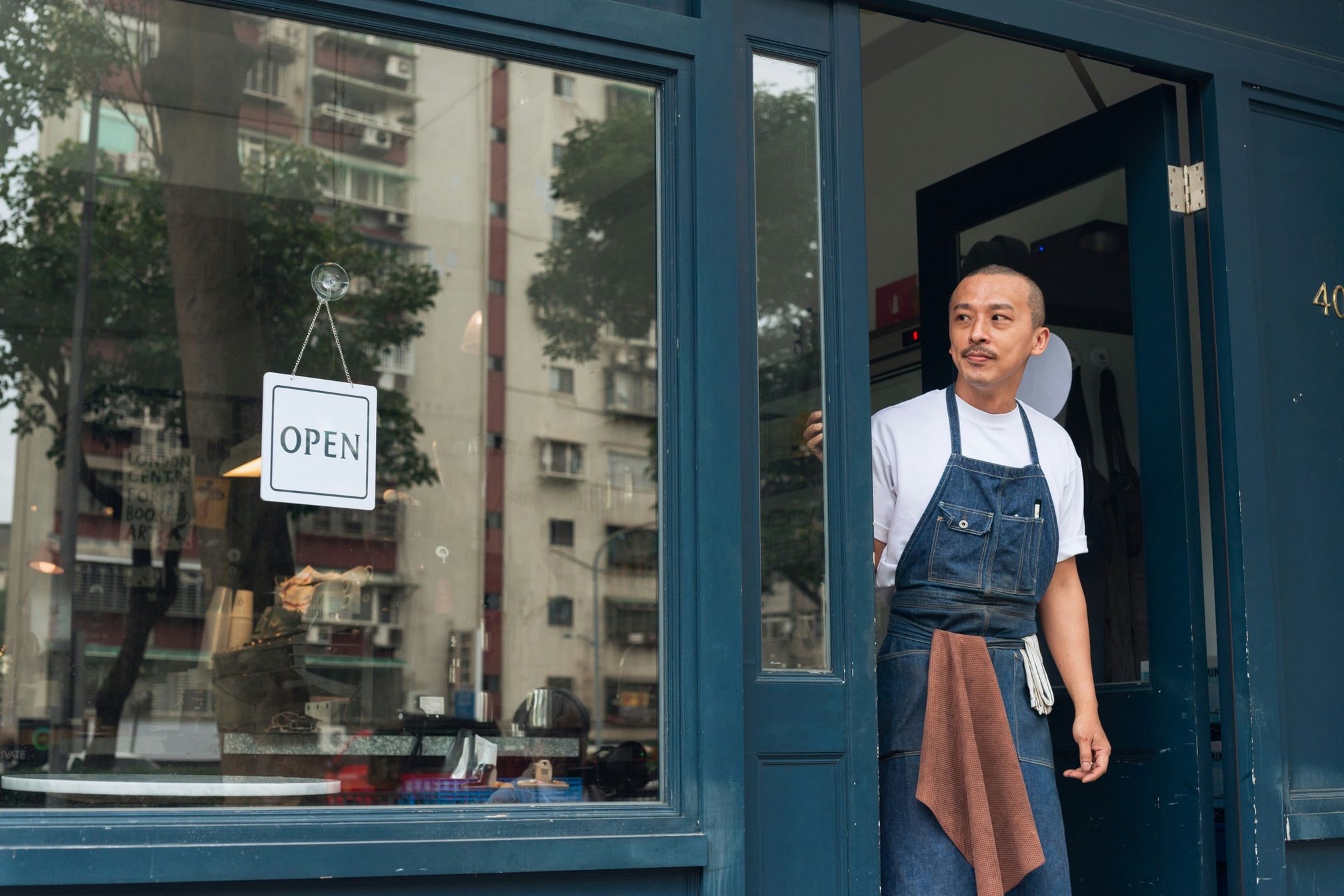 An apron-wearing worker stands in a blue storefront doorway beside an OPEN sign.