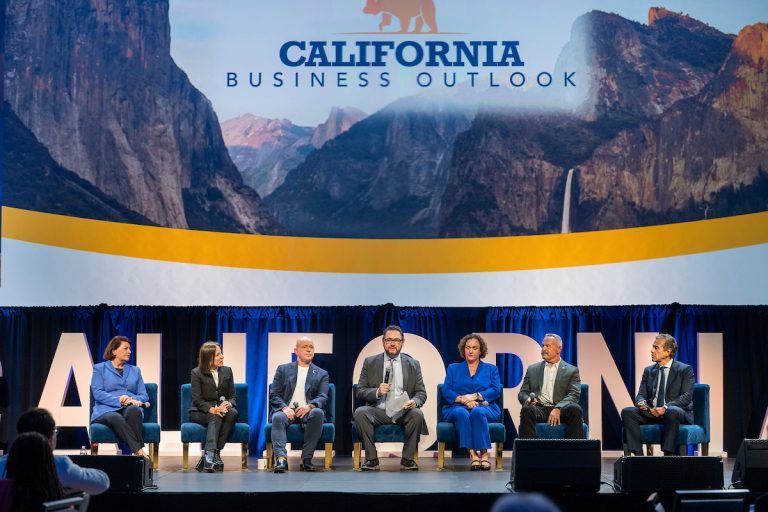 Seven panelists sit onstage beneath a California Business Outlook display featuring mountain scenery.